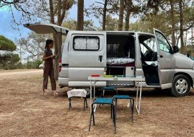 A woman prepares a meal outside a campervan in a forest setting in Portugal, highlighting the outdoor lifestyle offered by Atlantic Coast Campers.
